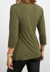 Maglietta aderente a maniche lunghe di colore verde oliva con scollatura rotonda e una texture liscia; abbinata a pantaloni neri. Orologio visibile sul polso sinistro.