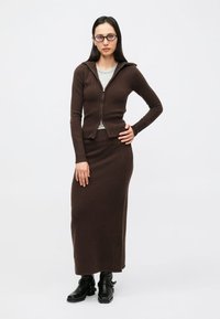 Barna kötött cipzáras kardigán szürke felső felett, barna hosszú szoknyával. Az összeállítás testhez simuló, bordázott textúrájú, fekete bokacsizmával párosítva.