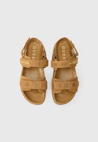 Καφέ σουέντ σανδάλια με δύο ρυθμιζόμενους ιμάντες, διακοσμητικά σε χρυσή απόχρωση με λογότυπο και επίπεδη σόλα. Διαθέτουν μαξιλαρωτό πέλμα για άνεση.