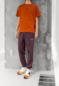 Rustoranžové bavlněné tričko s malým černým logem, spárované s fialovými tepláky s logem na kapsičce a bílými botami s oranžovými akcenty.