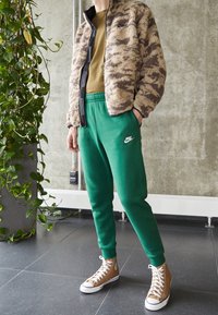 Tan och brun fuzzy jacka med svart dragkedja, olivgrön T-shirt, gröna sweatpants med vitt Nike-logotyp, och tan högskor.