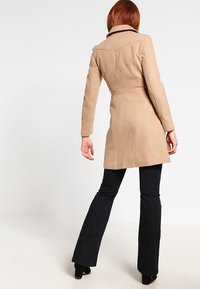Abrigo de mezcla de lana color beige con cuello estructurado, costuras ajustadas y cintura entallada, combinado con pantalones acampanados negros.