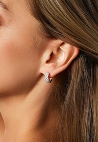 Nahaufnahme eines Ohres mit einem Creolenohrring, der mit kleinen funkelnden Steinen besetzt ist, und leichten braunen Haaren, die hinter dem Ohr gesteckt sind.