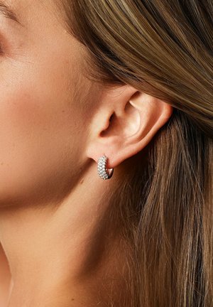 Nærbillede af øre med en hoops øresmykke indsat med små funklende sten og lysebrunt hår, der er stukket bag øret.