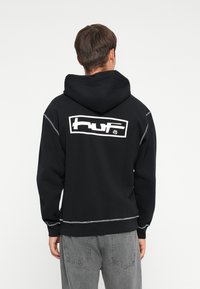 Svart hettegenser med en stor hvit "HUF" logo på baksiden, med mansjettstrikkede ermer og justerbar hette; mykt materiale, minimalistisk design.