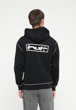 Black hoodie with a large white "HUF" logo on the back, featuring cuffed sleeves and a drawstring hood; soft material, minimalistic design.