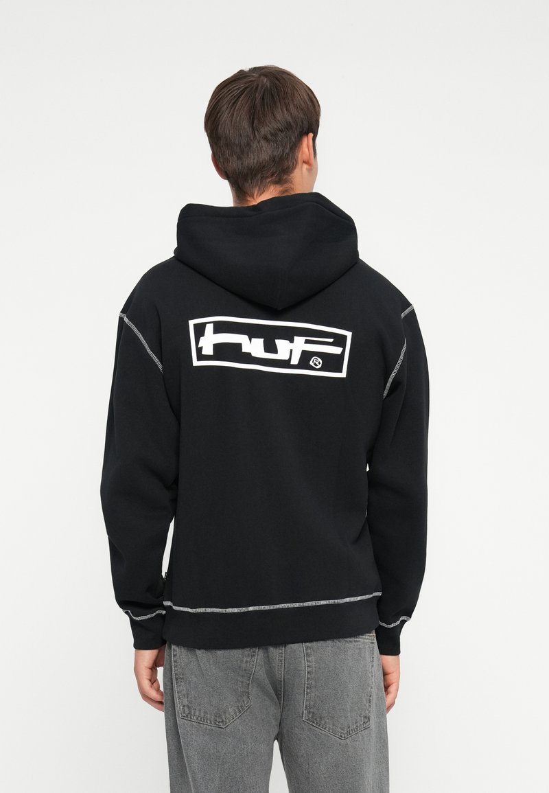 Svart hettegenser med en stor hvit "HUF" logo på baksiden, med mansjettstrikkede ermer og justerbar hette; mykt materiale, minimalistisk design.