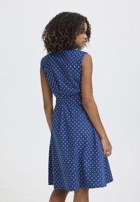 Žena s kučeravými vlasmi, oblečená v tmavomodrých šatách bez rukávov s bielymi bodkami, stojaca a pozerajúca sa nabok na jednoliatom pozadí.