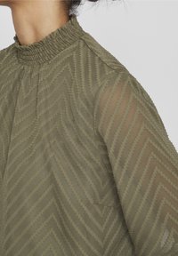 Blouse vert olive avec un col froncé haut, présentant un motif en zigzag texturé et des manches longues translucides.