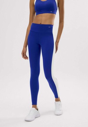 Mujer vistiendo leggings azules de cintura alta con paneles laterales blancos y un sujetador deportivo a juego, combinados con zapatillas deportivas blancas.