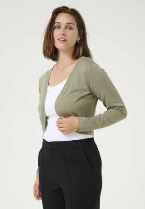 Olive green cropped cardigan, lightweight knit material, v-neck design, long sleeves, paired with a white top and black trousers.