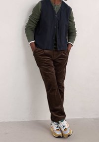 Chaleco azul marino oscuro sobre una camisa de manga larga verde oliva, combinado con pantalones de pana marrones y zapatillas blancas con acentos multicolores.