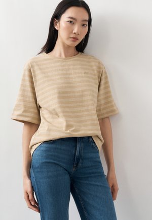 Ung kvinde med glat mørkt hår iført beige stribet T-shirt og blå jeans, stående mod en ensfarvet hvid væg.