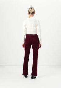 Vrouw met blond haar in een knot, gekleed in een witte kabelgebreide trui en bordeauxrode corduroy broek, staande tegen een effen witte muur.