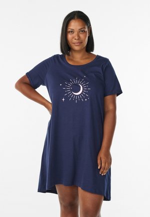 Frau mit kurzem, glattem Haar, die ein dunkelblaues Kleid mit einem Design aus Mondsicheln und Sternen trägt und mit einer Hand auf der Hüfte steht.