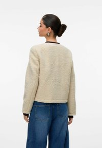 Krémová nadýchaná bunda s kratším střihem, kulatým výstřihem a žebrovanými manžetami, v kombinaci s modrými širokými džíny. Jednoduchý design, měkká textura.