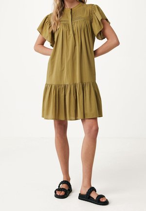Kobieta w oliwkowej sukience sięgającej do kolan z plisowanym gorsetem i falbaną na dole, w połączeniu z czarnymi sandałami na plecionej pasku.