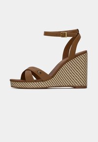 Sandal en cuir marron avec semelle compensée en tissu tissé diagonal. Dispose d'une sangle de cheville avec une boucle dorée.