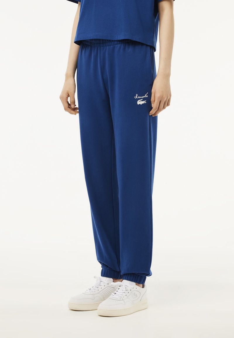 Blaue Jogginghosen aus weichem Material, mit einem elastischen Bund und Bündchen, sowie weißen bestickten Logo-Details an der Seite.