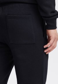 Juodi medvilniniai laisvalaikio kelnės su didžiule kišene nugaroje, rankogaliais ties čiurnomis ir lygiu paviršiumi, pavaizduotos iš šono su ranka ant jų.