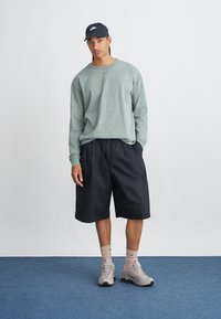 Hanorac verde deschis cu mânecă lungă, pantaloni scurți negri, adidași gri deschis și o șapcă închisă. Modelul stă pe un covor albastru, cu mâinile în buzunare.