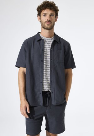 Kurzärmeliges Hemd mit Knöpfen in dunkelblau, strukturierter Stoff mit einer Brusttasche, darüber ein helles, gemustertes T-Shirt und passende Shorts.