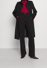 Cappotto nero con colletto alto, abbinato a un dolcevita rosso aderente, pantaloni neri a vita alta e tacchi neri a punta. Texture lisce in tutto.