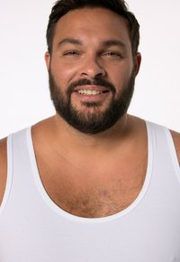 Hvid tanktop båret af en mand med en medium bygning, mørkt hår og skæg, som smiler mod en ensfarvet hvid baggrund.