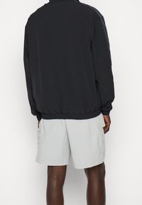 Person som bär en svart jacka med elastisk nederkant och manschetter, kombinerad med ljusgrå shorts med sidofickor, visad bakifrån.