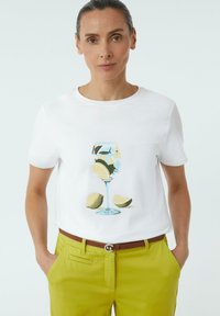 Weißes Baumwoll-T-Shirt mit einem Druck eines Glases Limonade und Zitronenscheiben. Trägt dazu leuchtend grüne Hosen und einen braunen Gürtel.