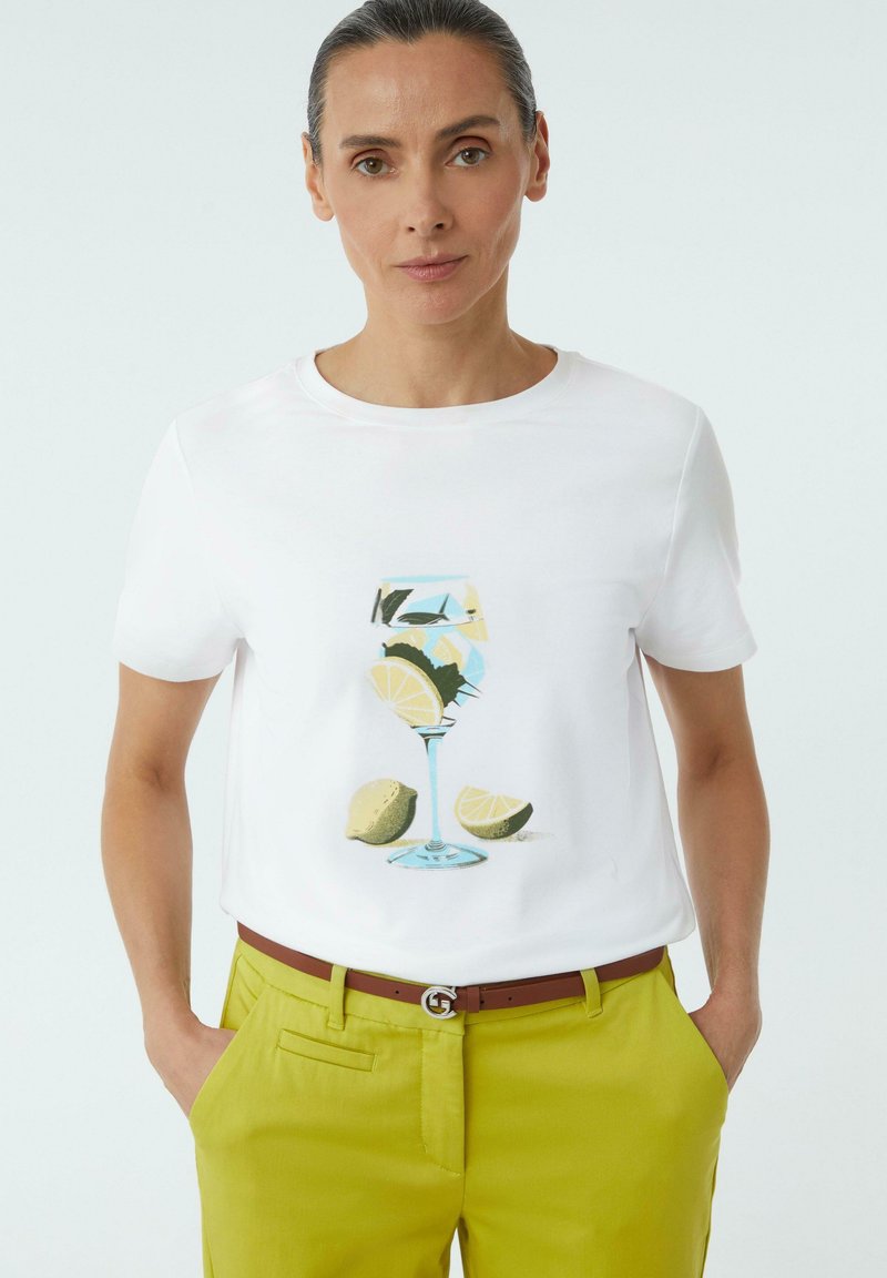 Weißes Baumwoll-T-Shirt mit einem Druck eines Glases Limonade und Zitronenscheiben. Trägt dazu leuchtend grüne Hosen und einen braunen Gürtel.