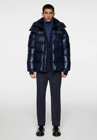 Mornarsko plava prozračna jakna s sjajnom, prešivenom teksturom, podesivim kapuljačom i prednjim patentnim zatvaračem. Nošena preko mornarsko plavog rolka i odijelnih hlača.