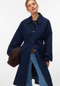 Γυναίκα που φοράει μπλε σκούρο παλτό με ζώνη, ανοιχτό μπλε τζιν και κρατάει ένα διπλωμένο σκούρο καφέ σακάκι μπροστά σε απλό λευκό φόντο.