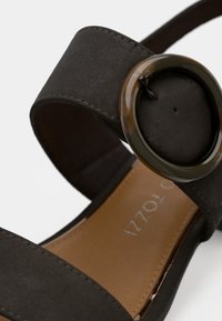 Μαύρη σουέτ σανδάλι με φαρδύ λουρί και κυκλικό πράσινο διακοσμητικό στο πλάι. Λείος καφέ πάτος και ραφές στα λουριά.