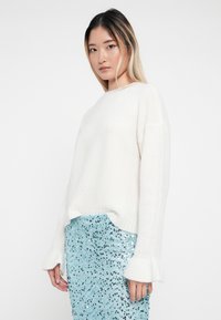 Weißer gestrickter Pullover mit Rundhalsausschnitt und glockenförmigen Ärmeln, kombiniert mit einem schimmernden blauen Paillettenrock.