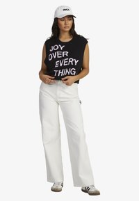 Black sleeveless knit top with "JOY OVER EVERY THING" in light pink text, paired with white wide-leg pants and a white cap.