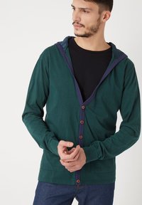 Sudadera de algodón verde con botones en la parte frontal, ribete en contraste de color marino y una capa interior negra. Presenta mangas largas y un ajuste casual.