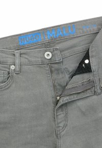 Harmaat farkut vetoketjukiinnityksellä, joissa on sininen "HUGO | MALU" -merkki vyötäröllä ja hopeisia koristereunoja. Tummat sisätaskut näkyvissä.