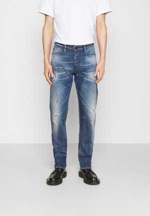 Man draagt verweerde blauwe jeans, wit T-shirt en zwarte leren schoenen, staand tegen een effen lichte achtergrond.