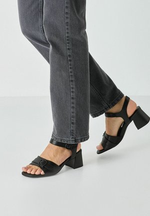 Black heeled sandals featuring a wide strap with a buckle, open toe, and a block heel. Worn with gray jeans, showcasing a casual look.