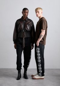Braune Leder-Cropped-Jacke mit Fronttaschen, schwarze Jogginghosen mit weißem Text und Schuhe, die Beige- und Brauntöne kombinieren.
