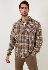 Camicia di flanella a quadri marrone e bianca con chiusura a bottoni, colletto e maniche lunghe, indossata con pantaloni beige su uno sfondo neutro.