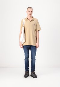 Beige polo skjorte med krage og liten logo, kombinert med blå jeans og svarte teksturerte joggesko. Holder en lyse farget totebag.