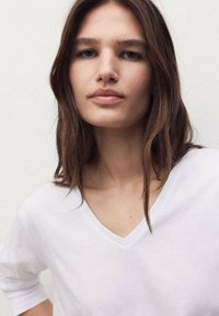 T-shirt bianca a V con maniche corte, dalle texture morbida e vestibilità rilassata, abbinata a capelli naturali e lisci e trucco minimale.