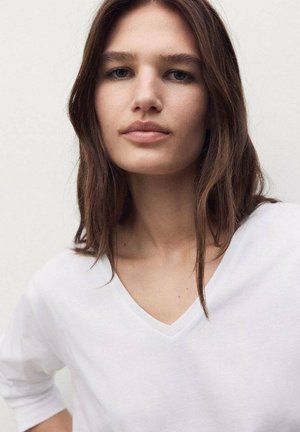 T-shirt bianca a V con maniche corte, dalle texture morbida e vestibilità rilassata, abbinata a capelli naturali e lisci e trucco minimale.