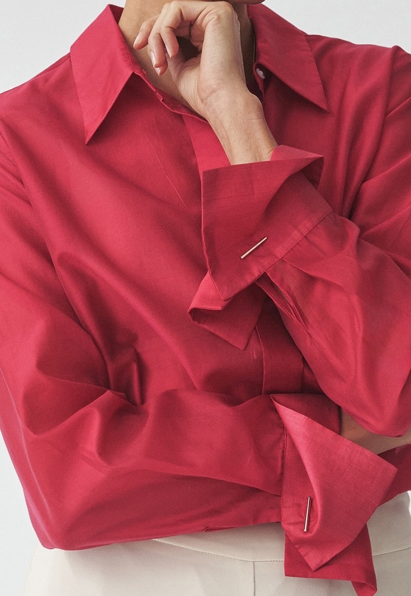 Personne portant une chemise rouge vif à manches longues avec des boutons de manchette, main reposant sur le menton, et pantalon blanc visible à la taille.