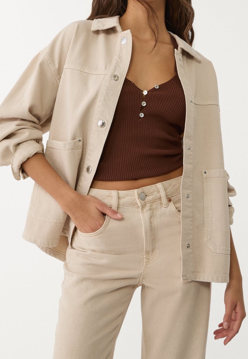 Beige denim-takki, jossa on hopeiset napit, yhden rinnan taskun ja käärittyjen hihojen kanssa, yhdistetty ruskeaan ribattuun paita ja vaaleanbeigeihin housuihin.