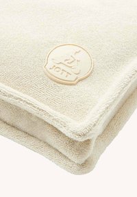 Toalla de felpa beige con superficie texturizada, con un logo circular JOTT color crema en relieve en una esquina.