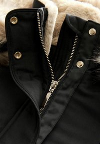Veste en duvet noire avec un col en fausse fourrure, quincaillerie dorée, fermeture éclair visible et boutons-pression, mettant en avant une texture douce.