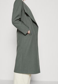 Grön ullblandad kappa med draperad front, stora slag och sidofickor. Har en rak silhuett och en något texturerad yta.